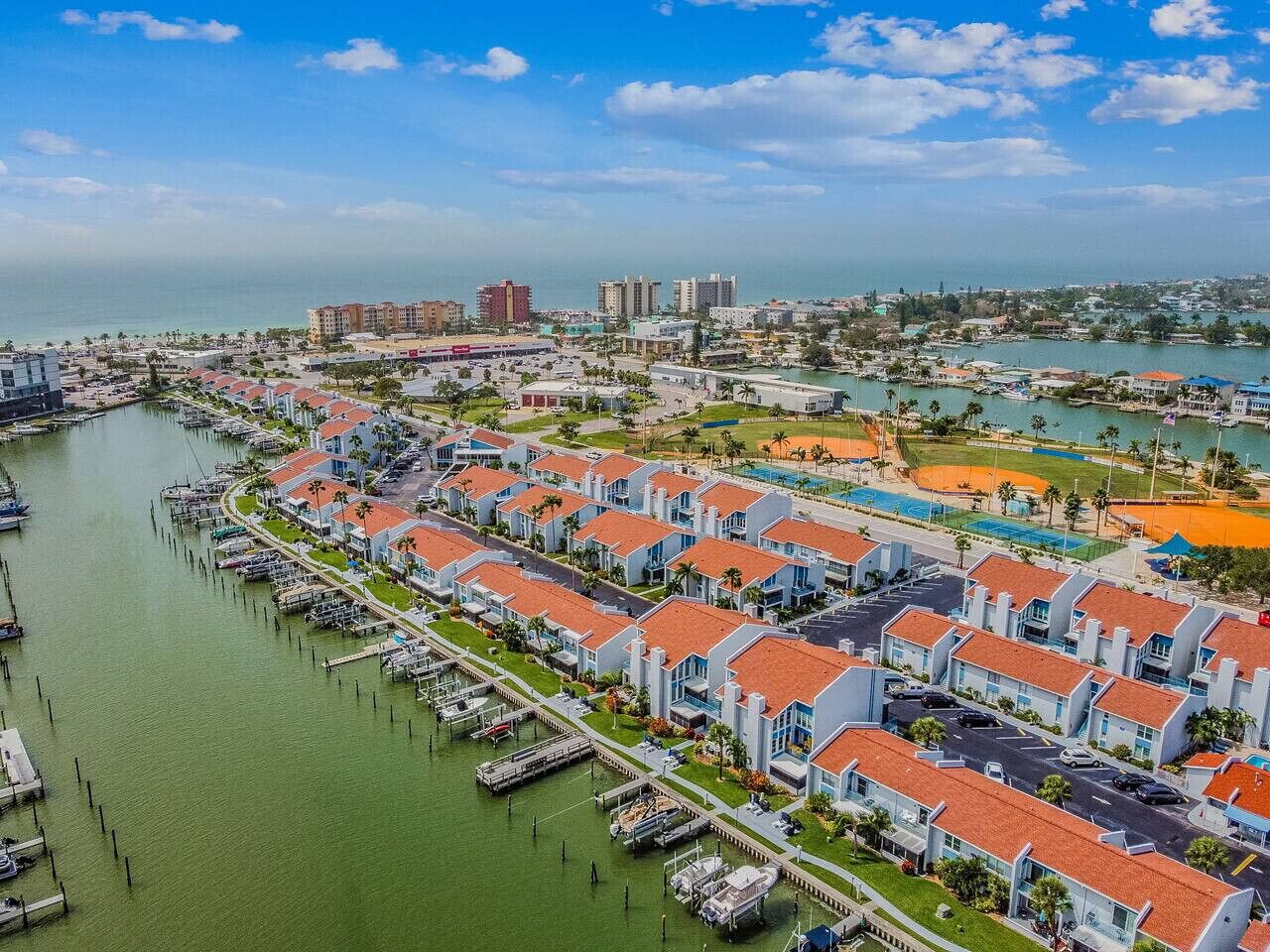 Una marina con barcos amarrados, una fila de edificios residenciales con tejados rojos, y una vista distante de un paisaje urbano con edificios y una masa de agua.