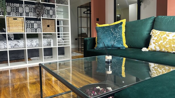 A modern living room with a glass coffee table, a green sofa with decorative pillows, a white shelving unit with baskets and textiles, and a mirror on the wall. - Comfortable 1-bed Chester Apartment with Parking & Full Kitchen, Home from Home (Chester)