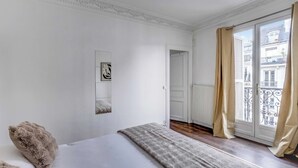 Un dormitorio con una cama, un ventanal con cortinas, un radiador y una puerta que da a otra habitación.