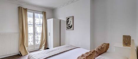 A bedroom with a bed, a window with curtains, a radiator, a wardrobe, and a wall-mounted artwork.