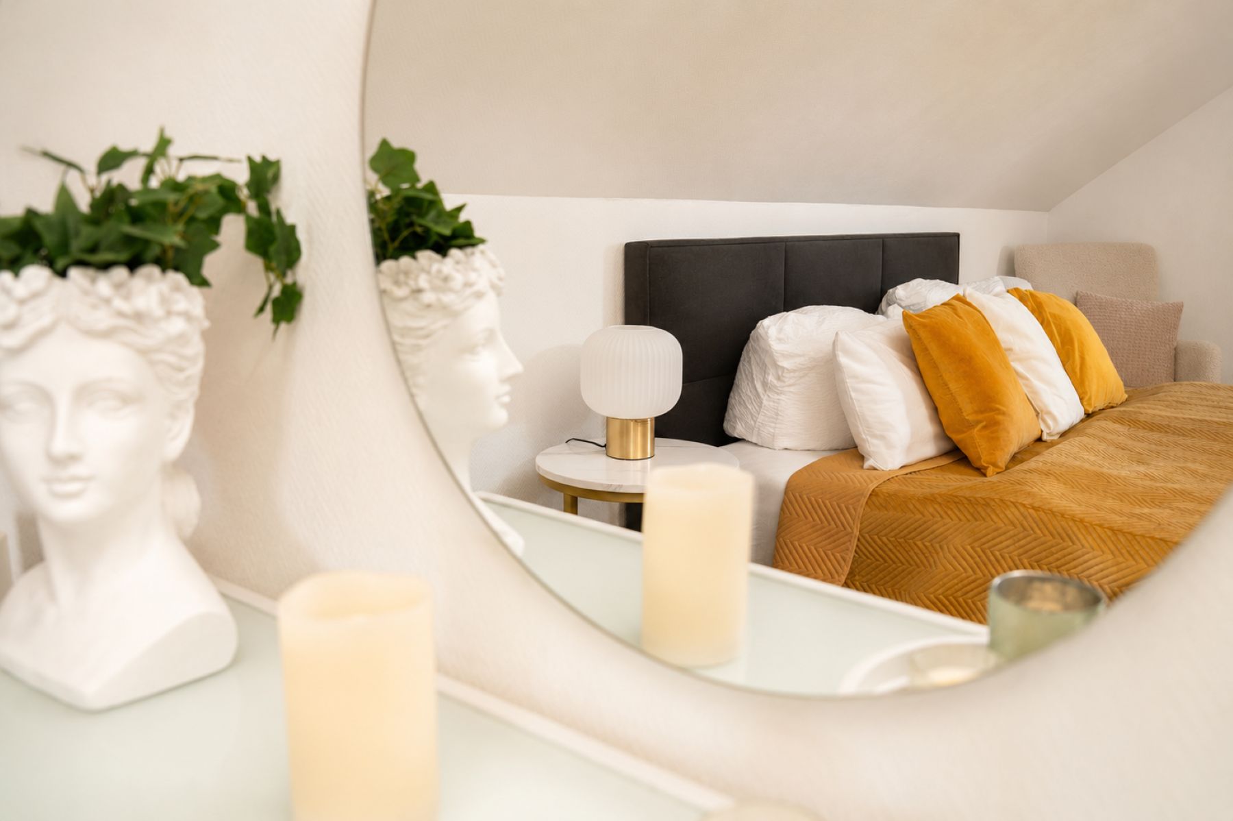 Ein Schlafzimmer mit Bett, Kissen, Nachttisch, Lampe und Spiegel, der den Raum widerspiegelt.