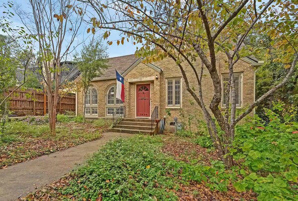 A brick building with a red door, a flag, and a small garden with a path leading to the entrance. - Park Free Walk to SoCo Ride to DT/UT/COTA (Austin)