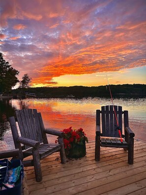 Two wooden chairs on a wooden dock with a fishing rod, a flower pot, and a calm lake reflecting the sunset.