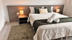 Ein ordentlich bezogenes Bett mit grauem Kopfteil, ein Nachttisch mit Lampe, eine grüne Tagesdecke und ein kleiner Blumentopf.
