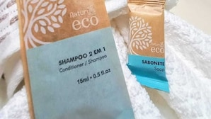 A bed with white linens, a blue pillow, and two eco-friendly shampoo and soap packages. - Pousada Aconchego em Mundaú (Trairi)