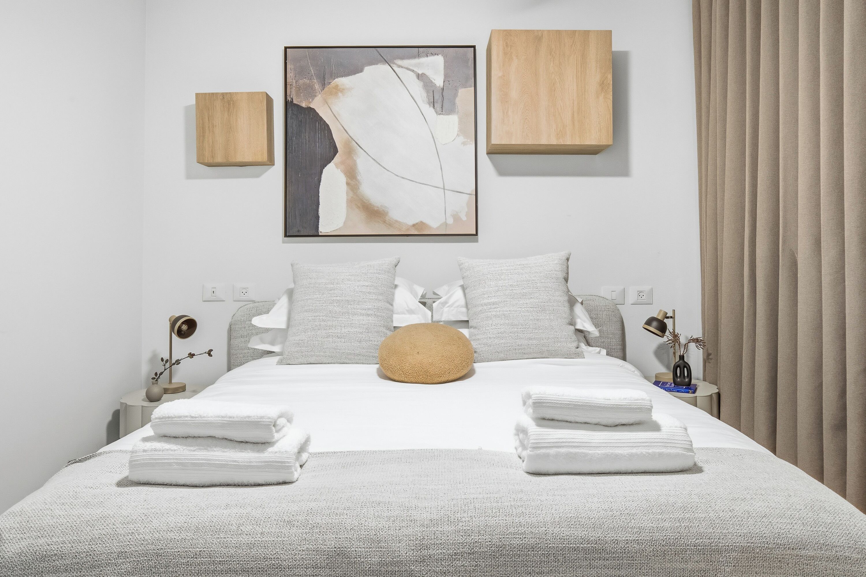 A neatly made bed with folded towels, a round cushion, and a painting above it. There are two wooden panels on the wall, a nightstand with a lamp, and a curtain on the window.