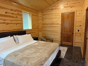 Une chambre à coucher située dans une cabane en bois, comprenant un lit, une table de chevet, une chaise, une fenêtre et une porte.