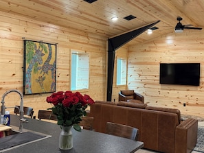 Un intérieur en bois comprenant un canapé, une table à manger avec des chaises, un vase de roses rouges, un tableau et un téléviseur fixé au mur.