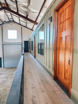 A long, narrow hallway with a wooden door on the right, a metal-framed window on the left, and a closed metal shutter door at the end. - Luxury Giraffe experience in our luxurious 4 bedroom solar powered Giraffe House (Lometa)
