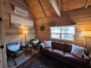 A cozy cabin interior with a brown leather sofa, two chairs, a small table with a plant, a mounted artwork, and a window with a view of trees.