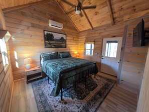 A wooden cabin bedroom with a bed, nightstand, lamp, air conditioning unit, and a door.