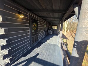 A covered wooden deck with a hanging chair, a wall-mounted light fixture, and a window with a wreath.