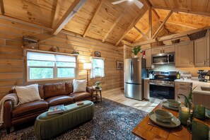 A cozy cabin interior with a wooden ceiling, a leather sofa, a dining table with dishes, and a kitchen area with appliances.