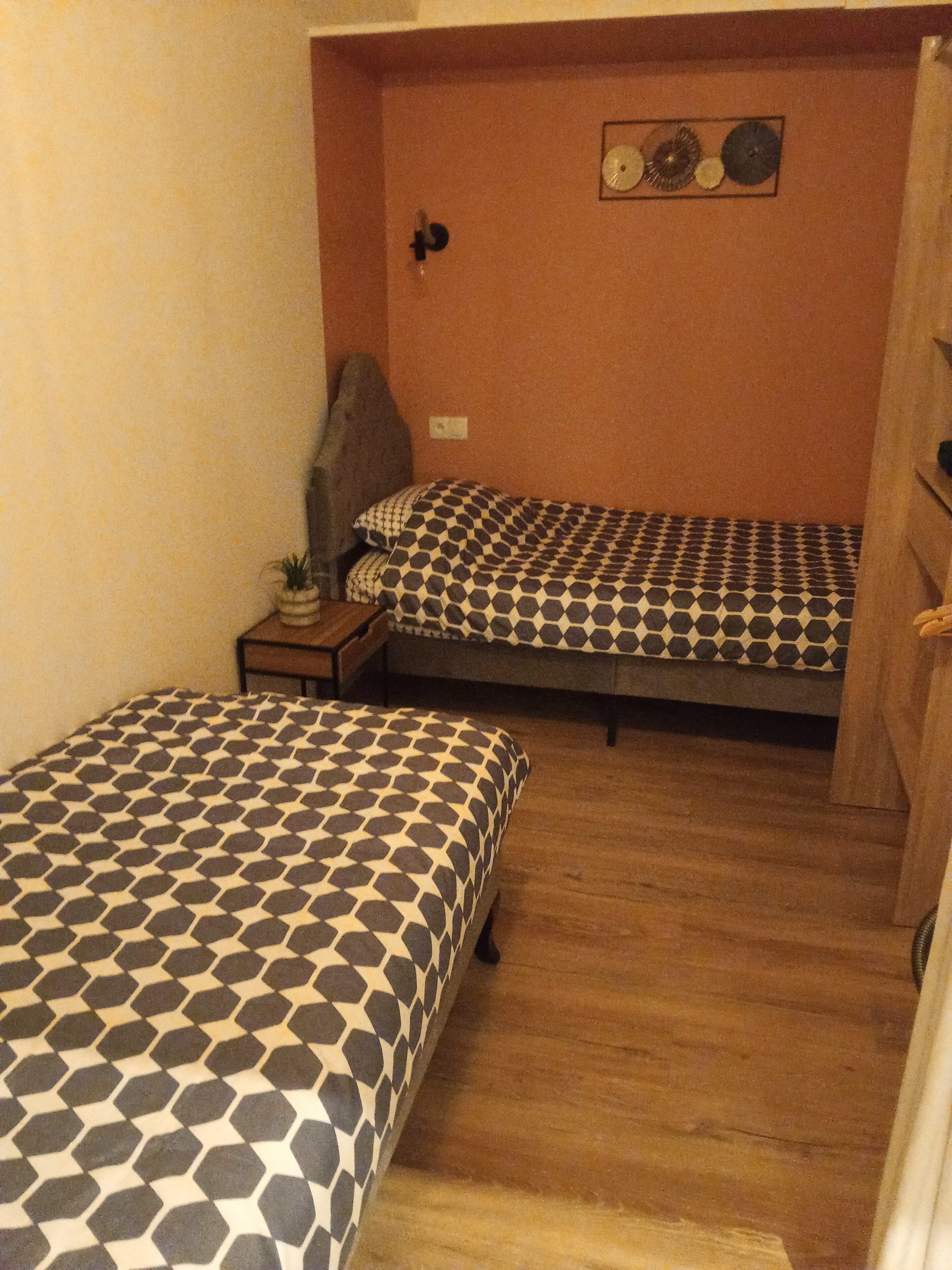 A small room with two beds, a wooden floor, a small table with a plant, and a wall-mounted clock.