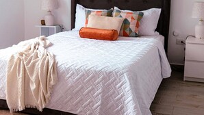 A neatly made bed with a white quilt, a brown throw blanket, and a variety of pillows. There are two bedside tables with lamps, a wall-mounted picture frame, and a wooden floor. - Beautiful, Modern and Central Apartment (Santo Domingo)