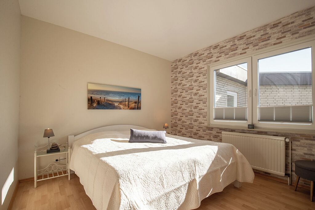 Un dormitorio con cama, mesita de noche, lámpara, un cuadro, ventana y pared de ladrillo.