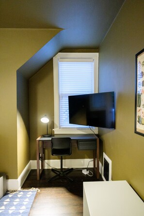 A room with a sloped ceiling, a desk with a chair, a flat-screen TV mounted on the wall, and a window with blinds.