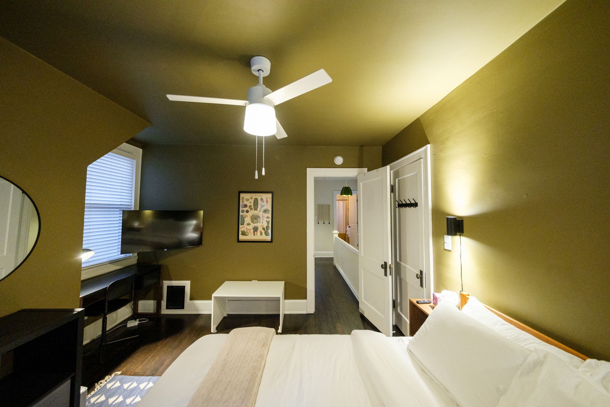 Ein Hotelzimmer mit einem Bett, einem Schreibtisch, einem Fernseher, einem Deckenventilator und einer Tür zu einem anderen Raum.