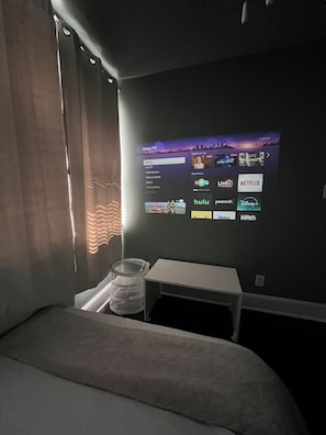 Hotellihuone, jossa on sänky, pieni pöytä ja seinälle asennettu näyttö, jossa näkyy television käyttöliittymä.
