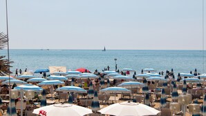 Une plage avec de nombreux parasols et des chaises, un ciel dégagé et un voilier à l’horizon.