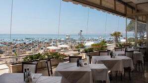 Une terrasse avec des tables et des chaises donnant sur une plage parsemée de parasols et de chaises longues.