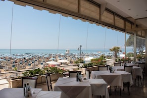 A terrace with tables and chairs overlooking a beach with umbrellas and lounge chairs. - Hotel Rivamare Jesolo (Jesolo)