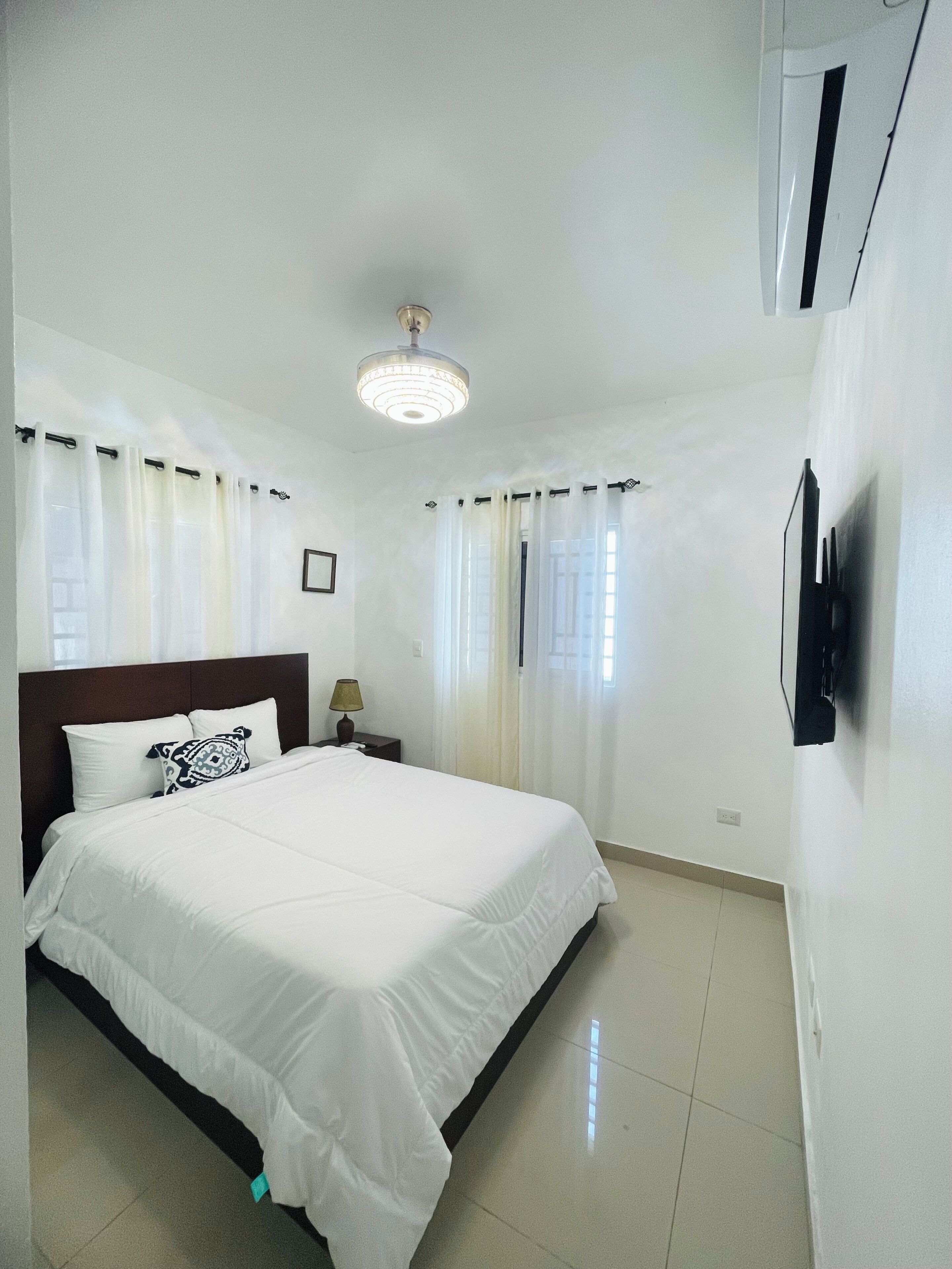 A hotel room with a bed, curtains, a nightstand, a lamp, a ceiling light, and an air conditioning unit.