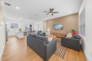 A spacious living area with a grey sofa, a coffee table, and a TV. There is a kitchen in the background with white cabinets and a dining area. - Ultimate Tampa Stay | Dual Units • 6BR • Sleeps 17 (Tampa)