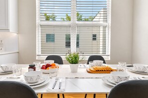 Mesa de comedor dispuesta con platos, tazones y vasos blancos, un centro de mesa con planta y fruta sobre la mesa.