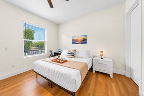A hotel room with a bed, a nightstand, a desk, a chair, a window with blinds, and a painting on the wall. - Ultimate Tampa Stay | Dual Units • 6BR • Sleeps 17 (Tampa)