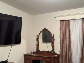 Ein Zimmer mit flachbildfernseher an der Wand, einer Holzkommode mit Spiegel und einem Fenster mit Vorhängen.