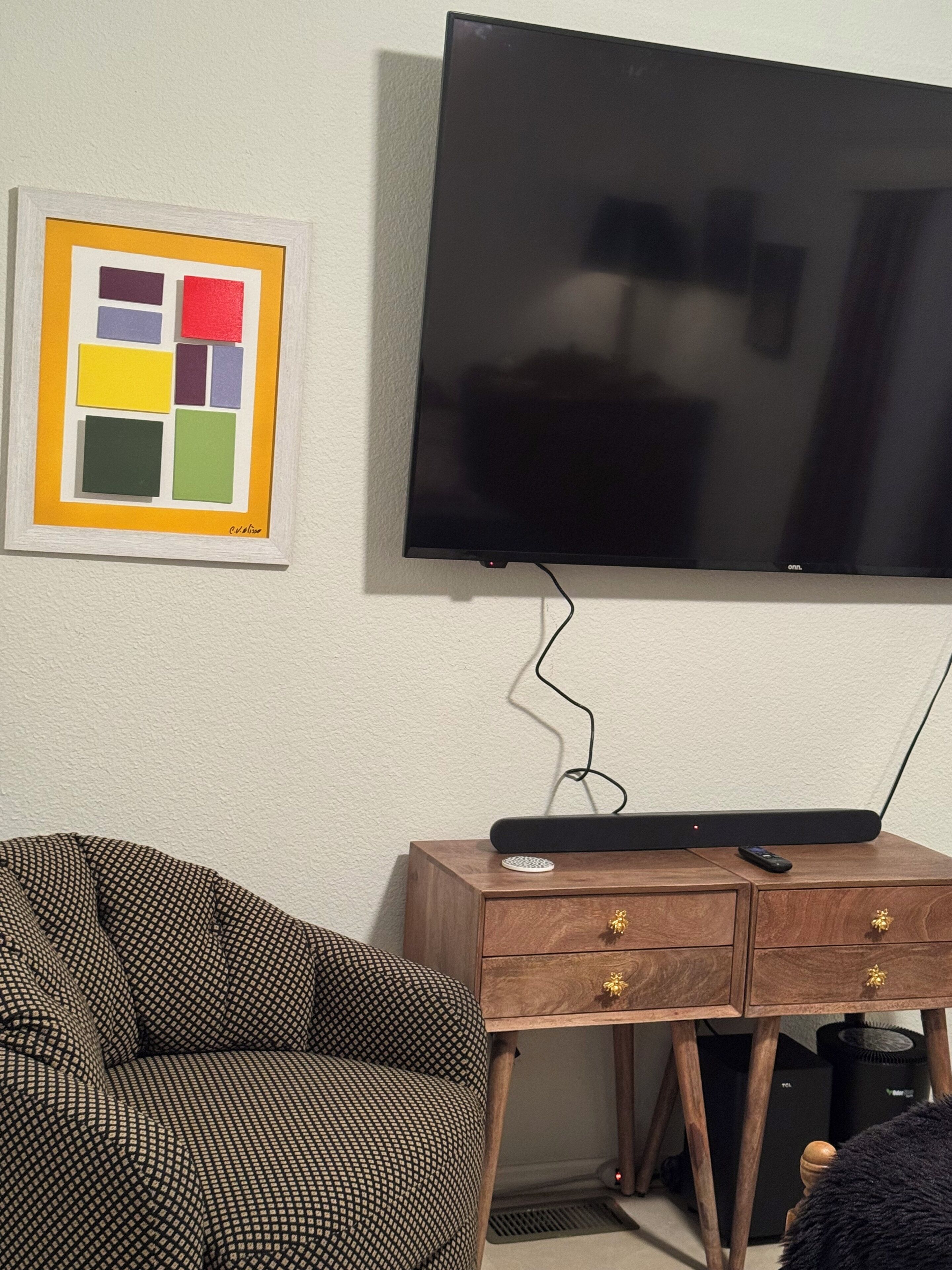 Quarto com televisão de tela plana na parede, sofá estampado, mesa de apoio de madeira com controle remoto e uma obra de arte emoldurada.