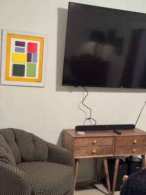Ein Zimmer mit einem an der Wand befestigten Flachbildfernseher, einem gemusterten Sofa, einem Holztisch mit Fernbedienung und einem gerahmten Kunstwerk.