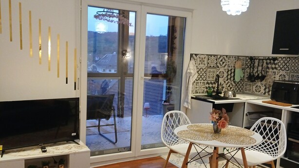 A modern living room with a flat-screen TV, white chairs, a patterned rug, and a kitchen area with a tiled backsplash.