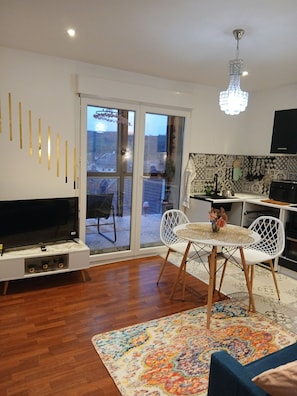 A modern living room with a flat-screen TV, white chairs, a patterned rug, and a kitchen area with a tiled backsplash.