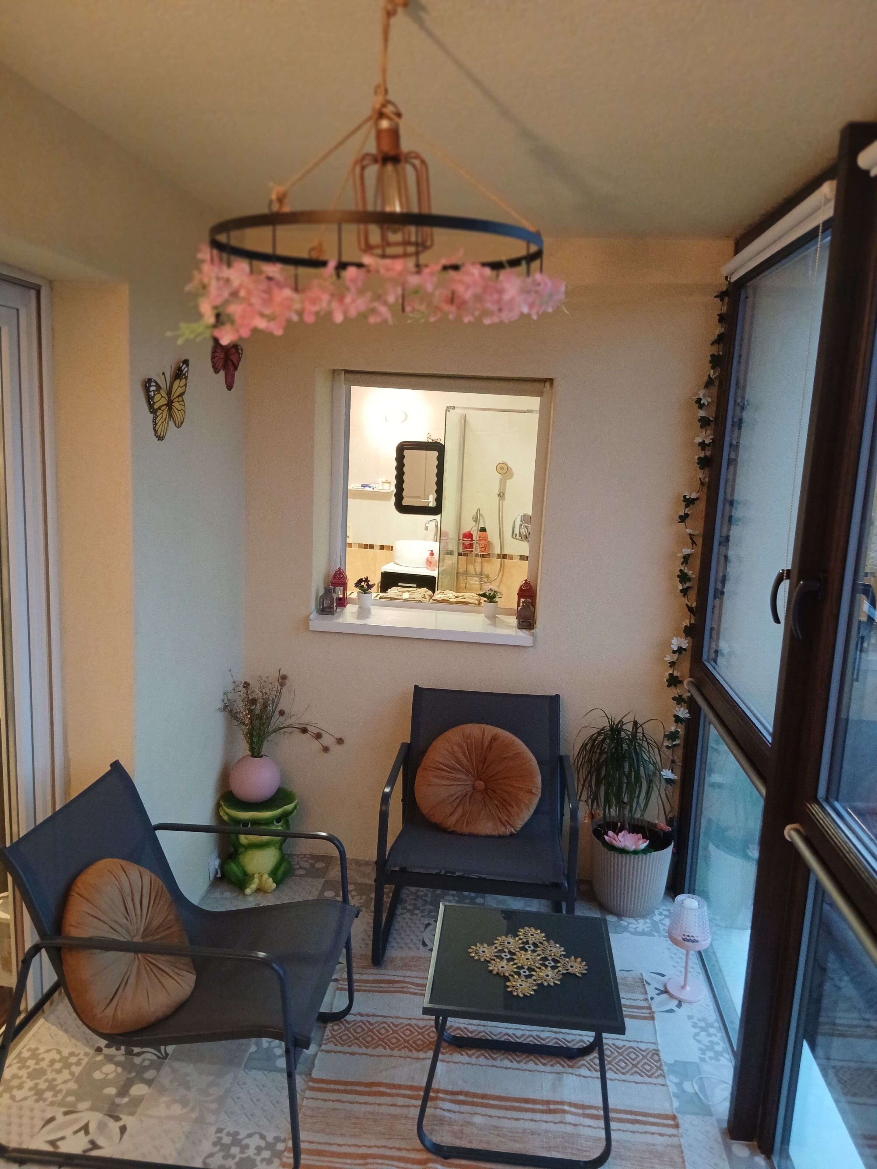 A small, cozy room with two chairs, a table, a hanging light fixture, a window with a mirror, and a potted plant.