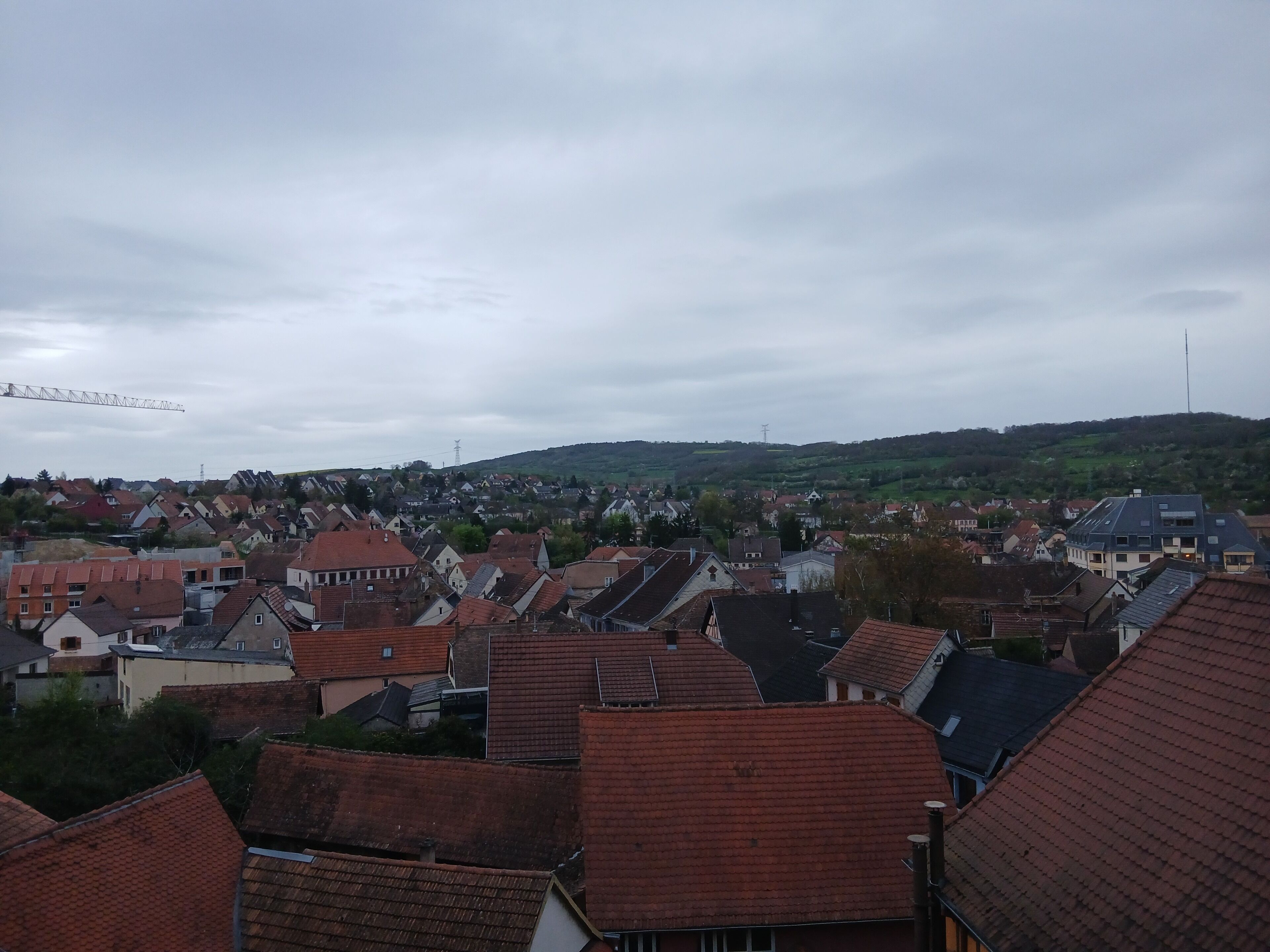 A residential area with houses featuring red-tiled roofs, a hill in the background, and a crane in the distance.