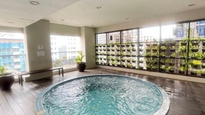 An indoor swimming pool with a tiled edge, surrounded by wooden flooring, large windows with a view of buildings, and a wall of hanging planters. - Cozy gated studio apartment in Central Jakarta with AC, fitness room, and pool (Jakarta Pusat)