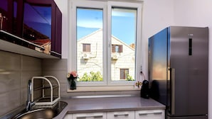 Una cocina con un lavamanos de acero inoxidable, armarios blancos, una ventana con vistas a edificios y un refrigerador.