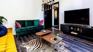 Una sala de estar con un sofá verde, un sofá amarillo, un tapete estampado con diseño de cebra, una mesa de centro, un televisor y una lámpara colgante original.