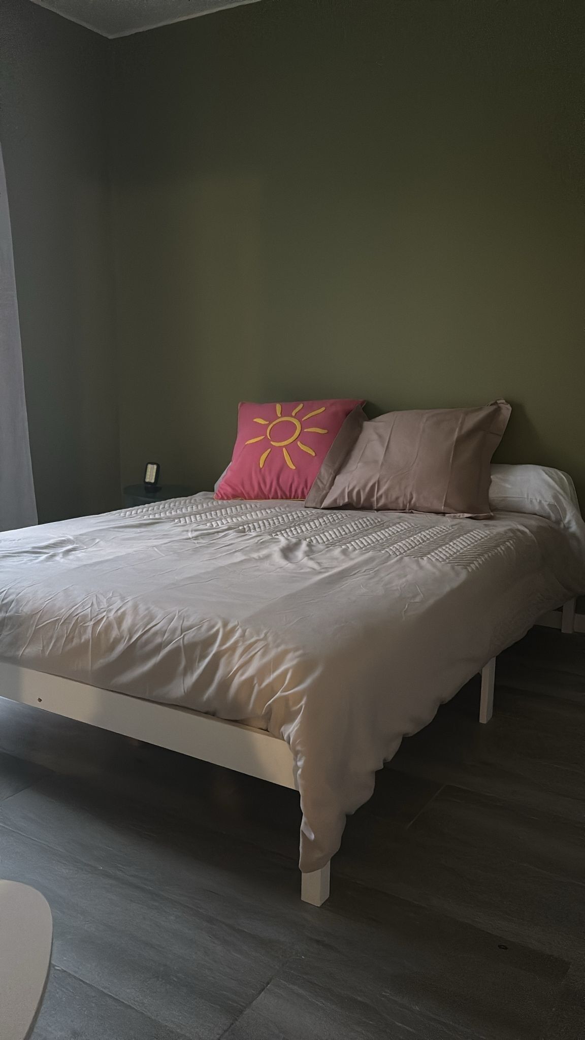A bed with a white frame, a grey mattress, and two pillows, one pink and one beige, in a room with a green wall and a wooden floor.