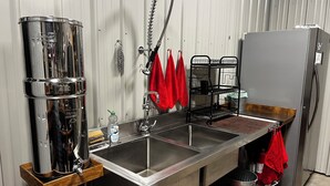 Una cocina de acero inoxidable con un lavamanos doble, un grifo colgante, un utensilero de madera, una rejilla metálica y un refrigerador gris.