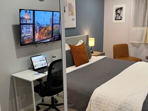 Ein Hotelzimmer mit Bett, Schreibtisch mit Laptop, Stuhl, Fernseher der eine Stadtansicht zeigt und einer Wand mit gerahmten Bildern.