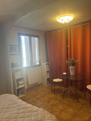 Une pièce comprenant un lit, une table avec des chaises, une plante en pot, une bibliothèque et une fenêtre avec des rideaux.