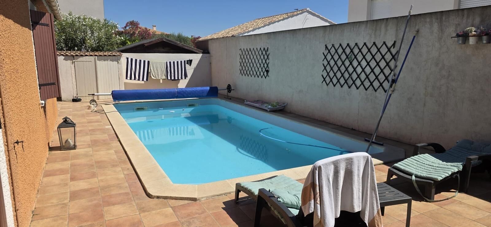 Et rektangulært swimmingpool med blå interiør, omgivet af en fliseterrasse. Liggestole og et lille bord med et håndklæde.