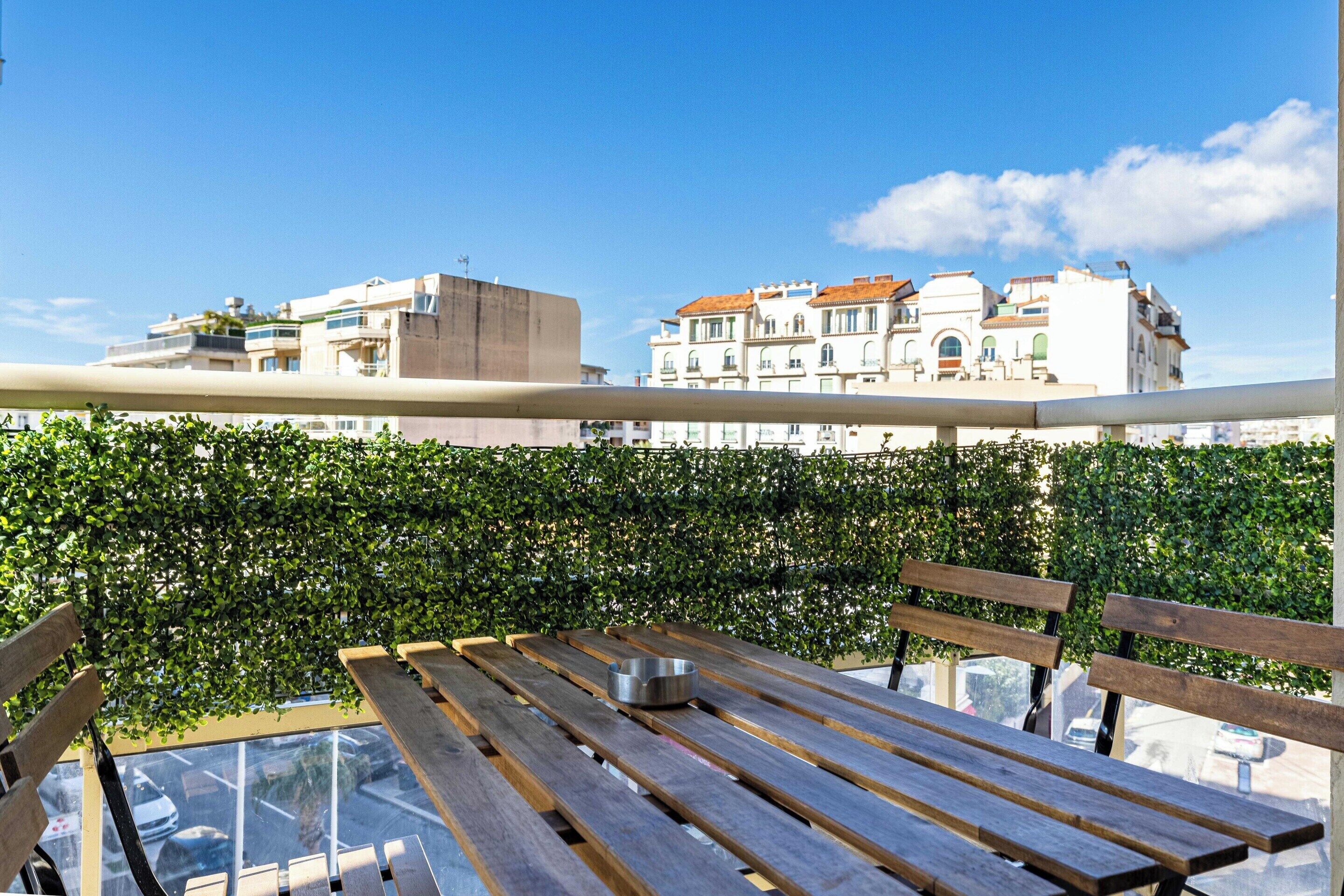 Un balcon avec une table et un banc en bois, un récipient en métal et une haie.