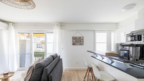 Een moderne keuken met een zwart aanrecht, witte barkrukken en een grijze bank in een lichte woonruimte met een houten vloer.