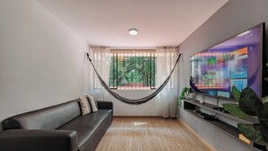 A living room with a black leather sofa, a hammock, a TV on a stand, and a window with a view of greenery. - Perfect 3-bedroom apartment in Medellín for a relaxing stay (Medellín)