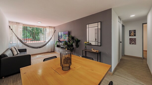 A modern living room with a hammock, a wooden dining table, a TV, and a large window with a view of greenery. - Perfect 3-bedroom apartment in Medellín for a relaxing stay (Medellín)