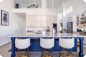 A modern kitchen with white cabinets, a blue island with three white bar stools, and a staircase leading to an upper level. - FIFA I 15 Mins to AT&T I 4BR I 5 Bed I 5.5 Bath I Sleeps 10 (irving)